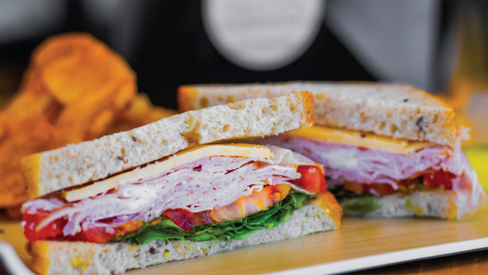 A ham and cheese sandwich with lettuce and tomato, cut in half, with chips on a plate