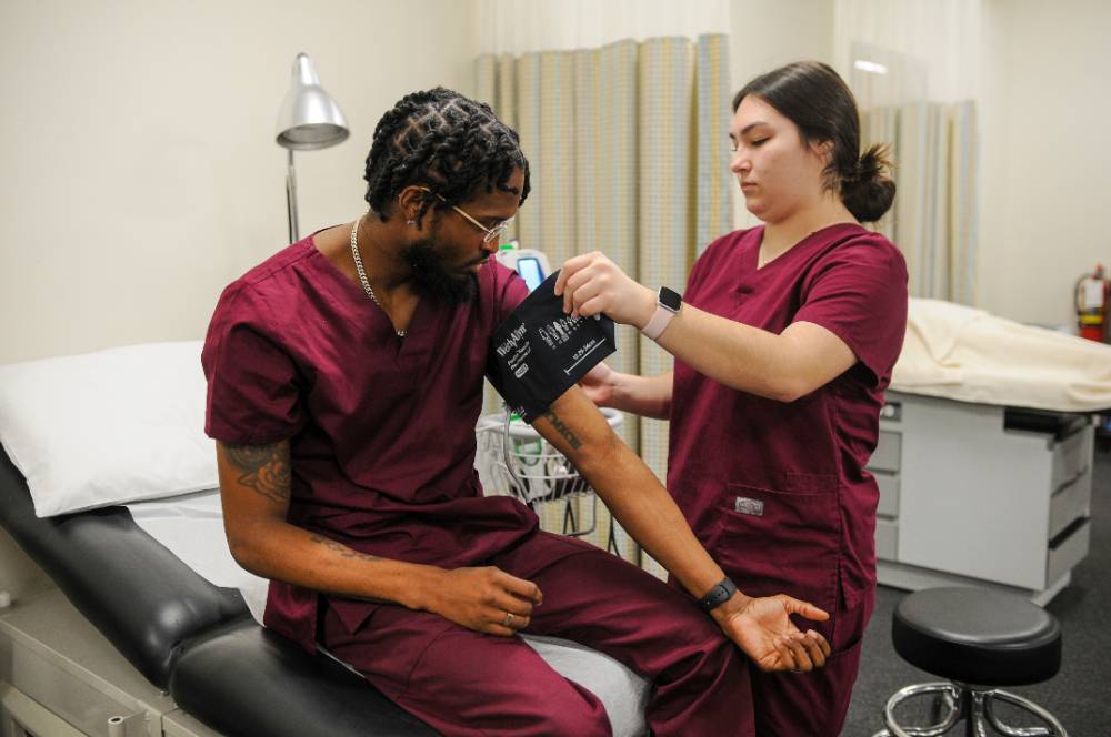 female student in maroon scrubs taking blood pressure of make student in maroon scrubs