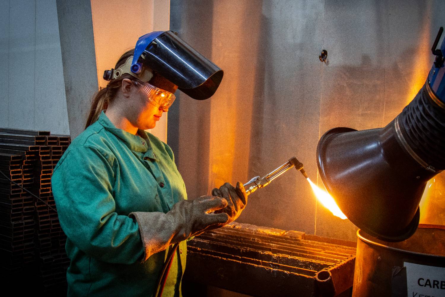Female welding student wearing protective gloves and green gear, the welding mask on her head with the shield flipped up, holding a lit torch