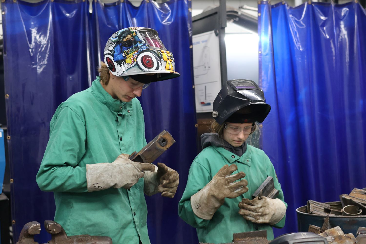 male and female welding students wearing protective gear, green smocks, gloves, and welding helmets with lids lifted up as they look down holding metal pieces