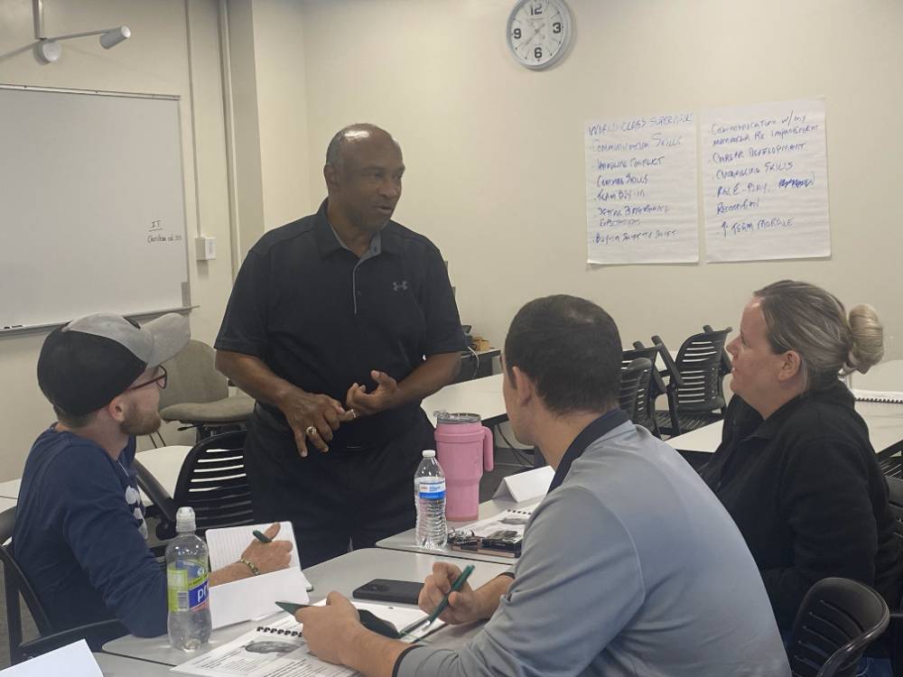 man standing among adult students who are sitting at a table and engaging in a conversation