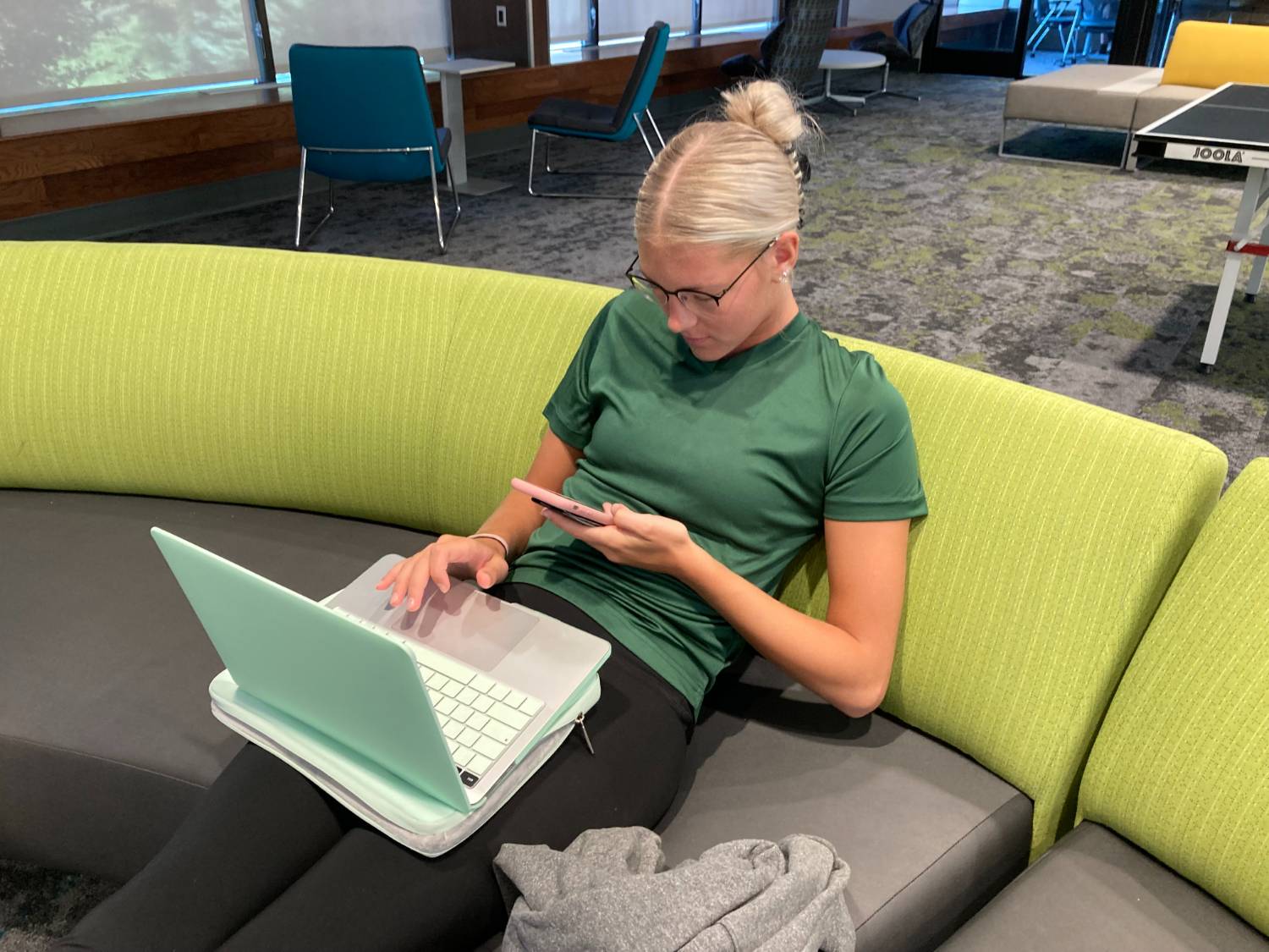 female student sitting on couch with laptop on her lap and looking at her phone