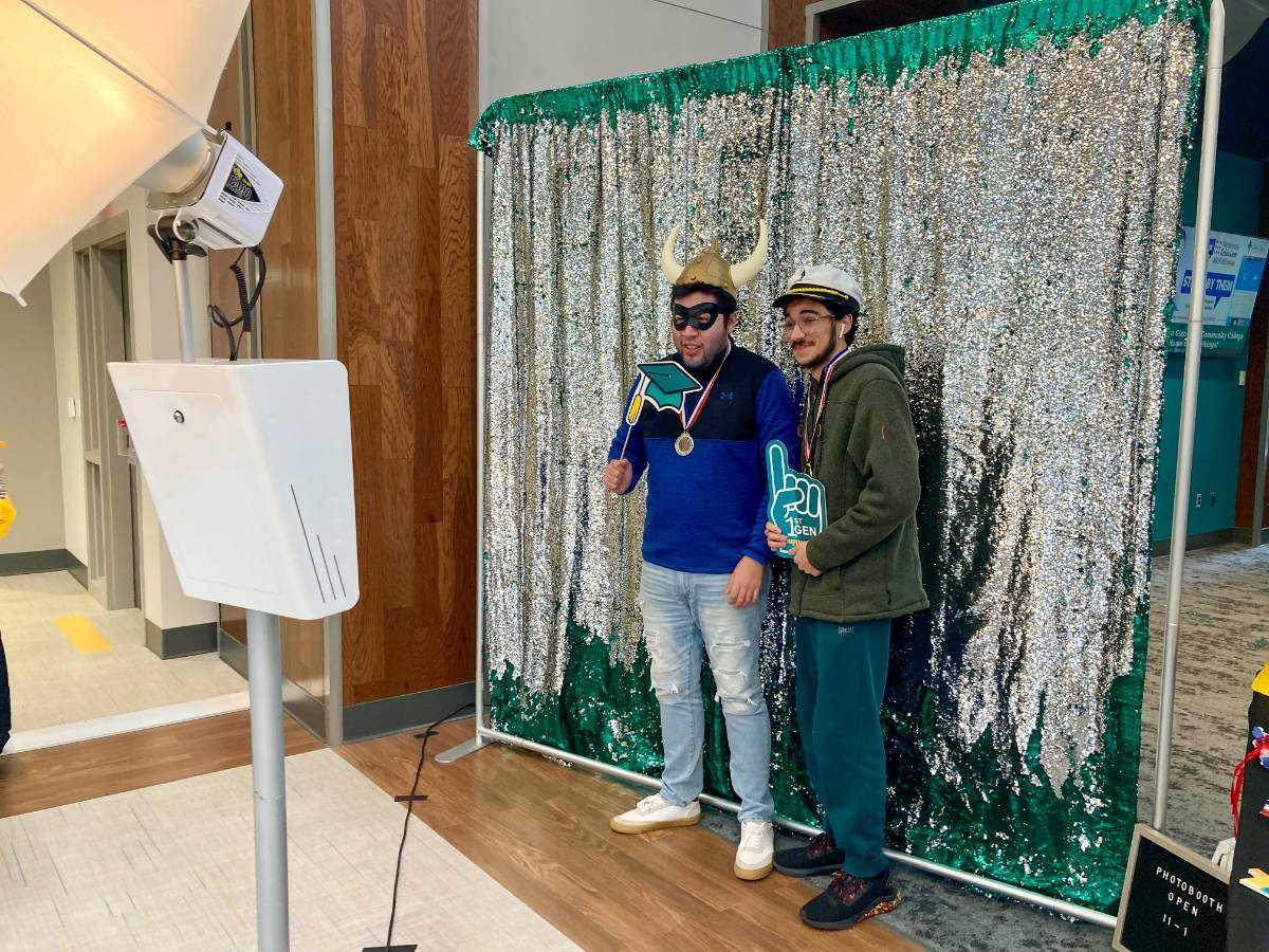 Two first-gen male students standing in front of a sequined backdrop, doing a photo shoot, wearing Viking helmets and holding first-gen props