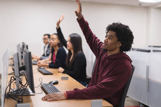 Students sit at workstations in the college computer lab. An African-American male is working on an assignment in the foreground. Several females are at the same table on different workstations. Two students are raising their hands to ask question.