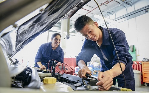 Two trainee car mechanics in garage repairing car, young man smiling and concentrating